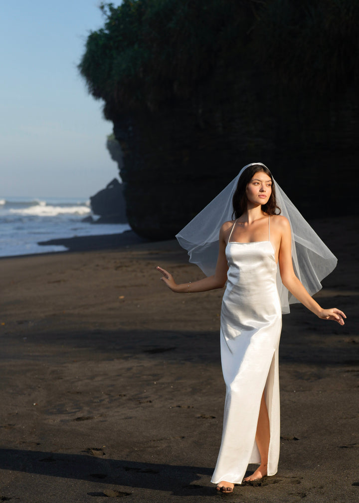 A bride in a white gown with a short simple plain classic veil, fingertip length, two tier standing on a beach with the ocean and cliffs in the background.