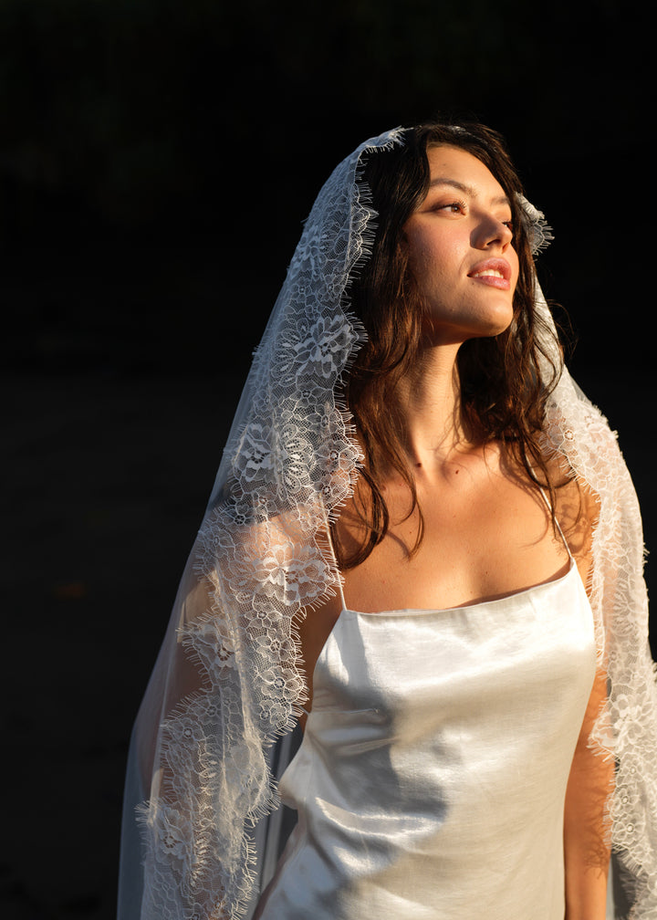 A woman wearing a white bridal gown and a mantilla chantilly lace veil, single tier with floral trim.