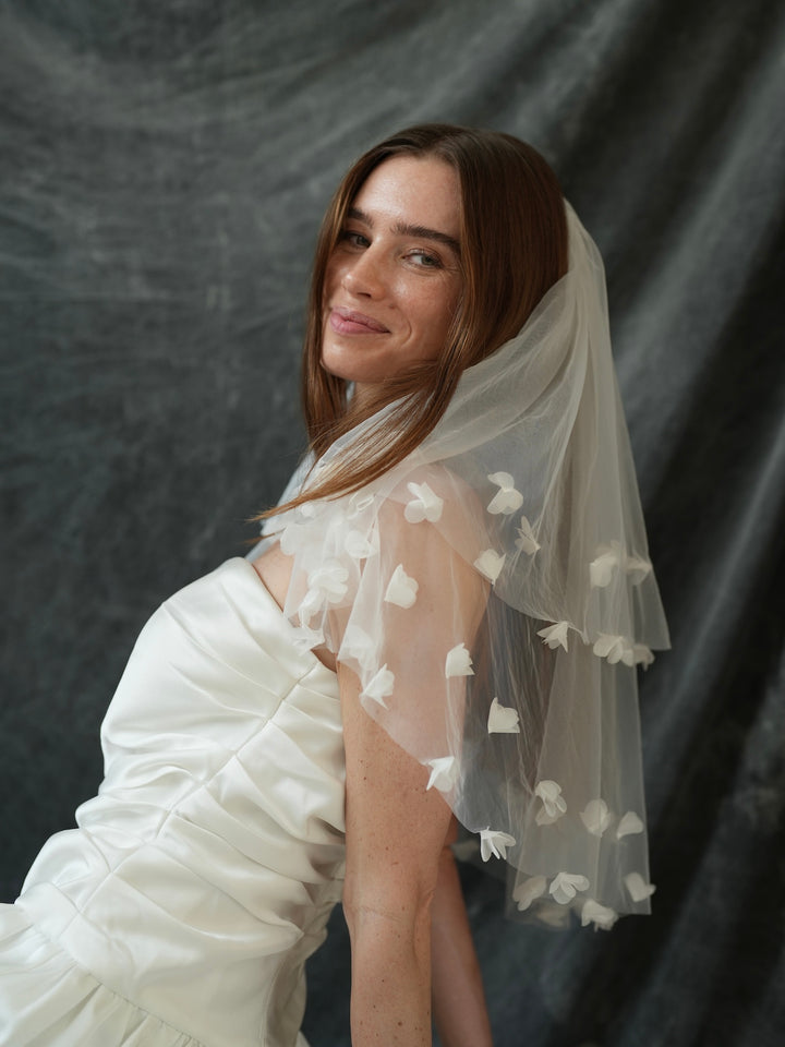 Woman wearing a white wedding dress with a mini 3d flower short two tier bachelorette veil against a dark background