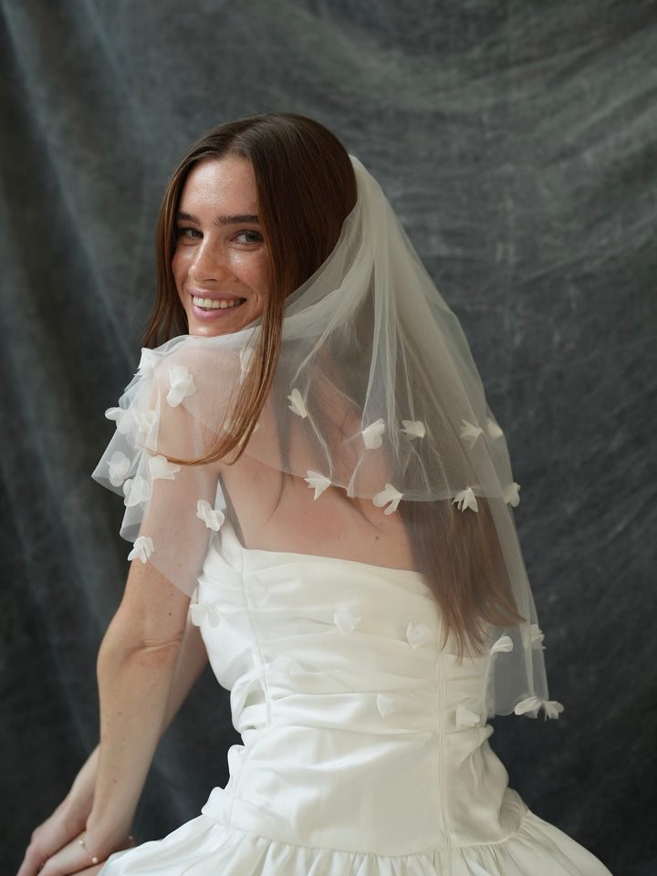 Woman wearing a white wedding dress with a 3d flower short two tier bachelorette veil against a dark background