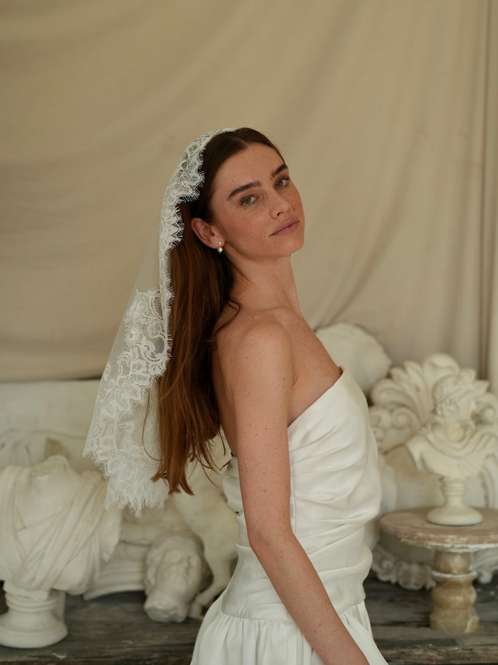 Woman in a white dress with a mantilla short lace bachelorette veil standing in front of classical sculptures.