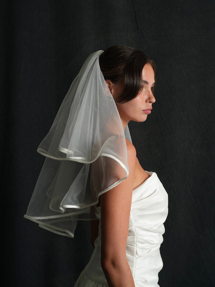 Woman wearing a mini satin edge short two tier bachelorette veil against a black background