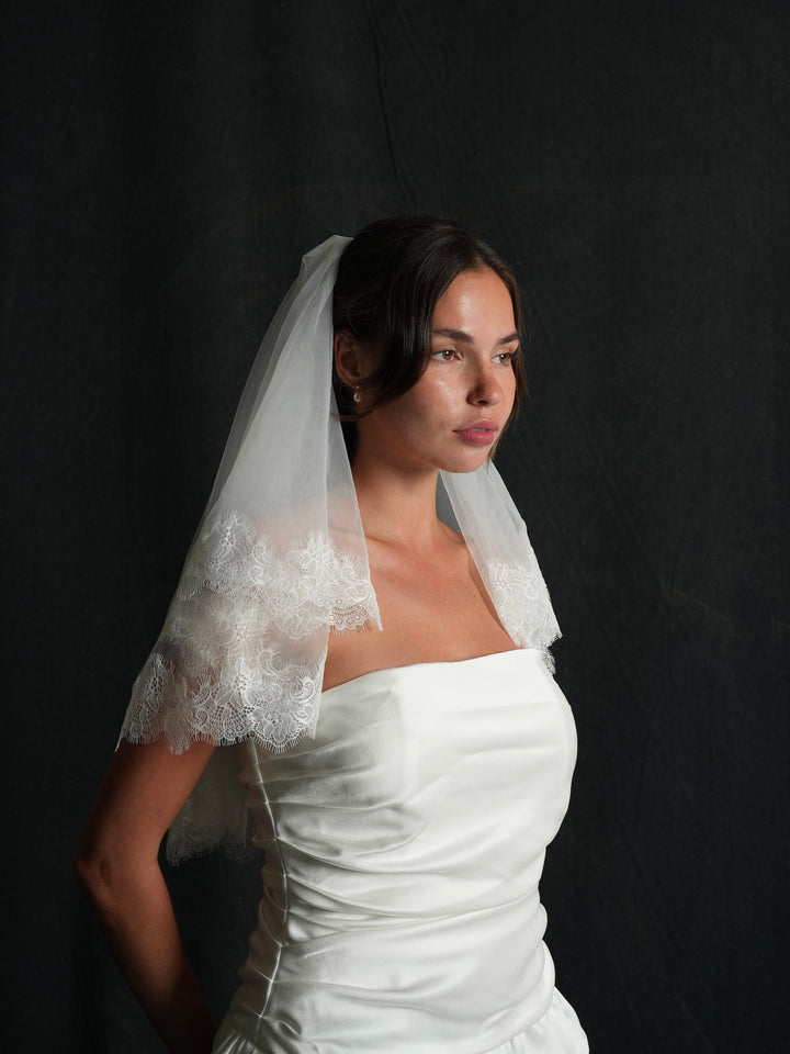 Woman wearing a white wedding dress with a mini chantilly lace short two tier bachelorette veil against a black background