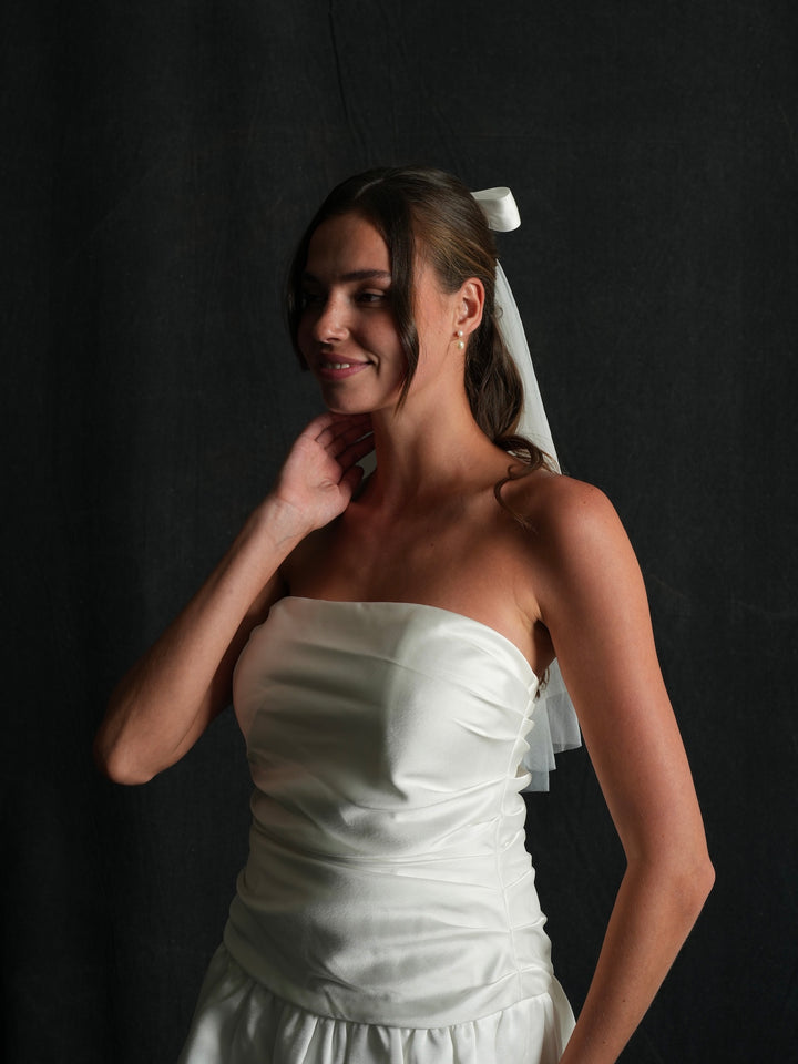Woman wearing a white strapless dress and mini bachelorette bow veil against a black background
