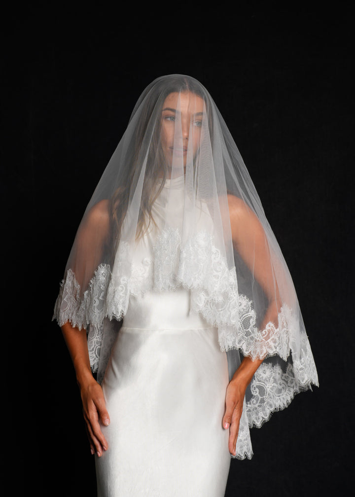 Bride wearing a white wedding dress with a short lace veil, two tier, fingertip length against a black background