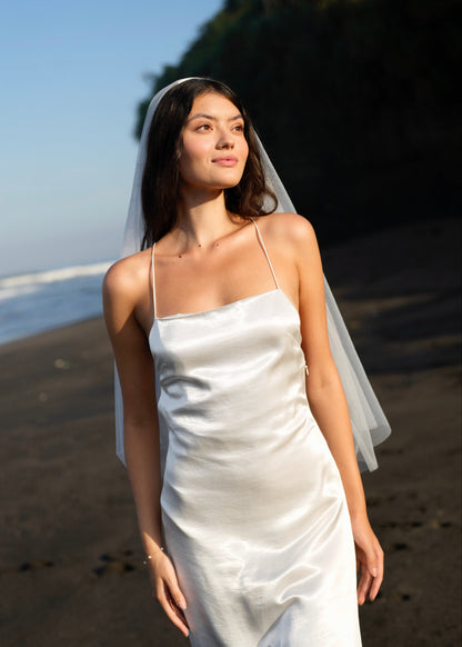 Woman in a white wedding dress with a short simple plain classic veil, standing on a beach.