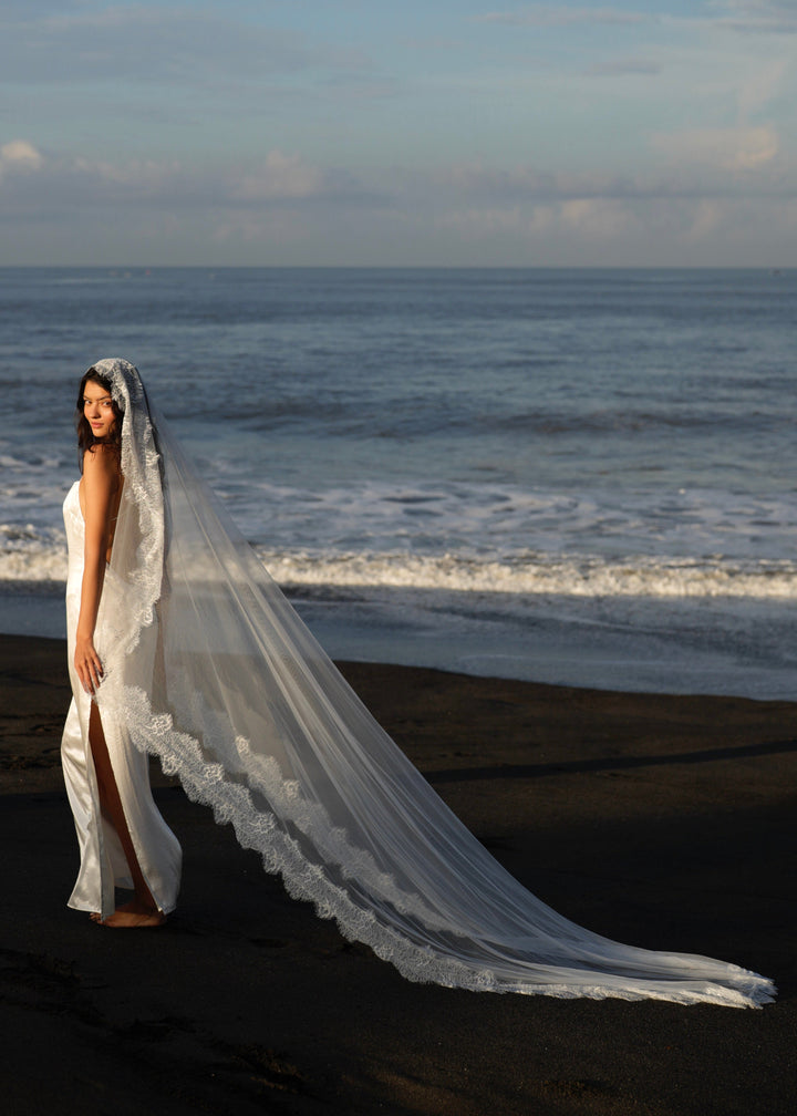 Woman in a white wedding dress with a mantilla chantilly lace veil, single tier, cathedral length standing on a beach.